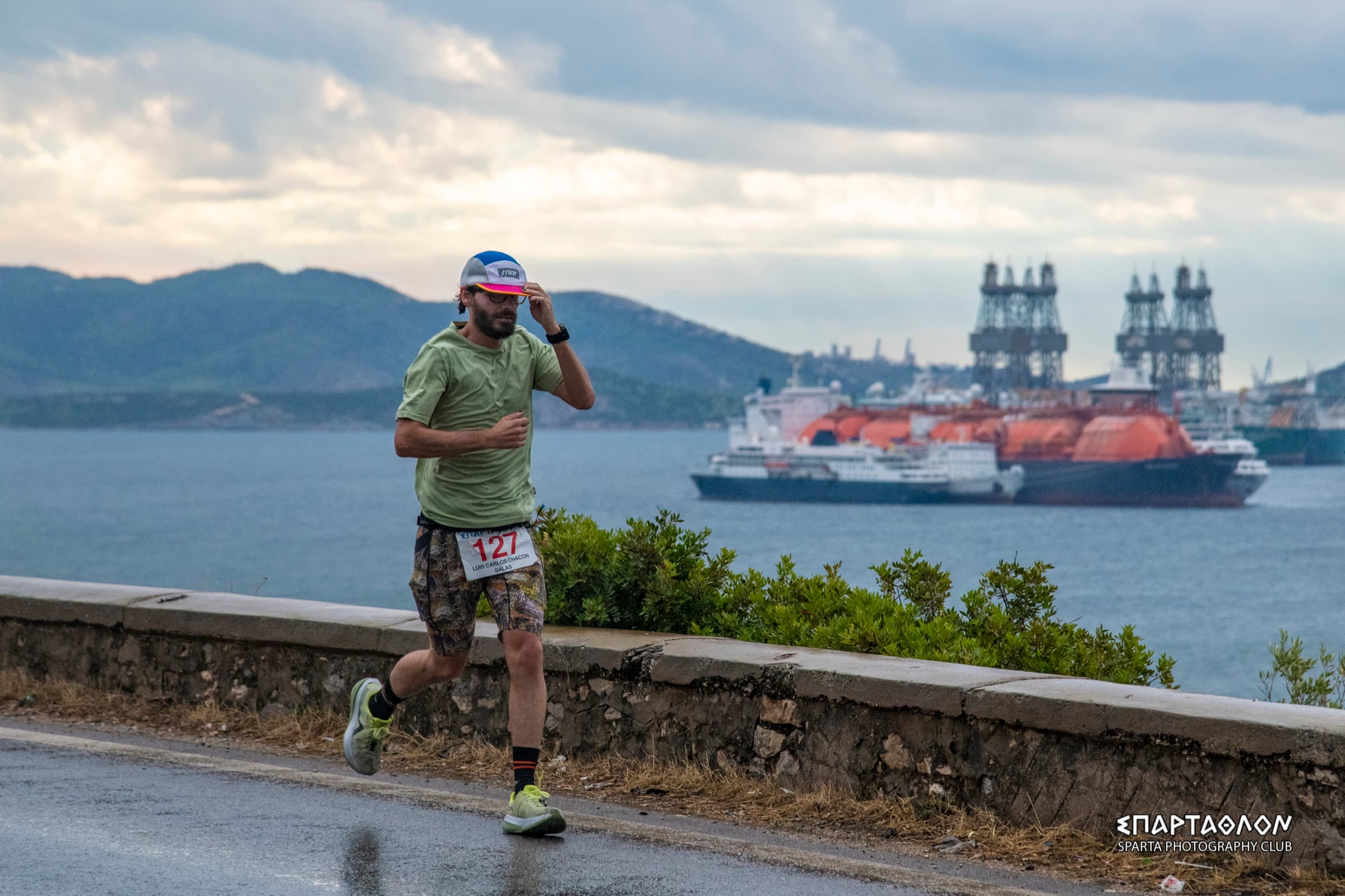 El autor corre por una carretera mojada junto al mar, con el dorsal 127. Detrás hay barcos grandes, grúas de un astillero y montañas bajo un cielo nublado.