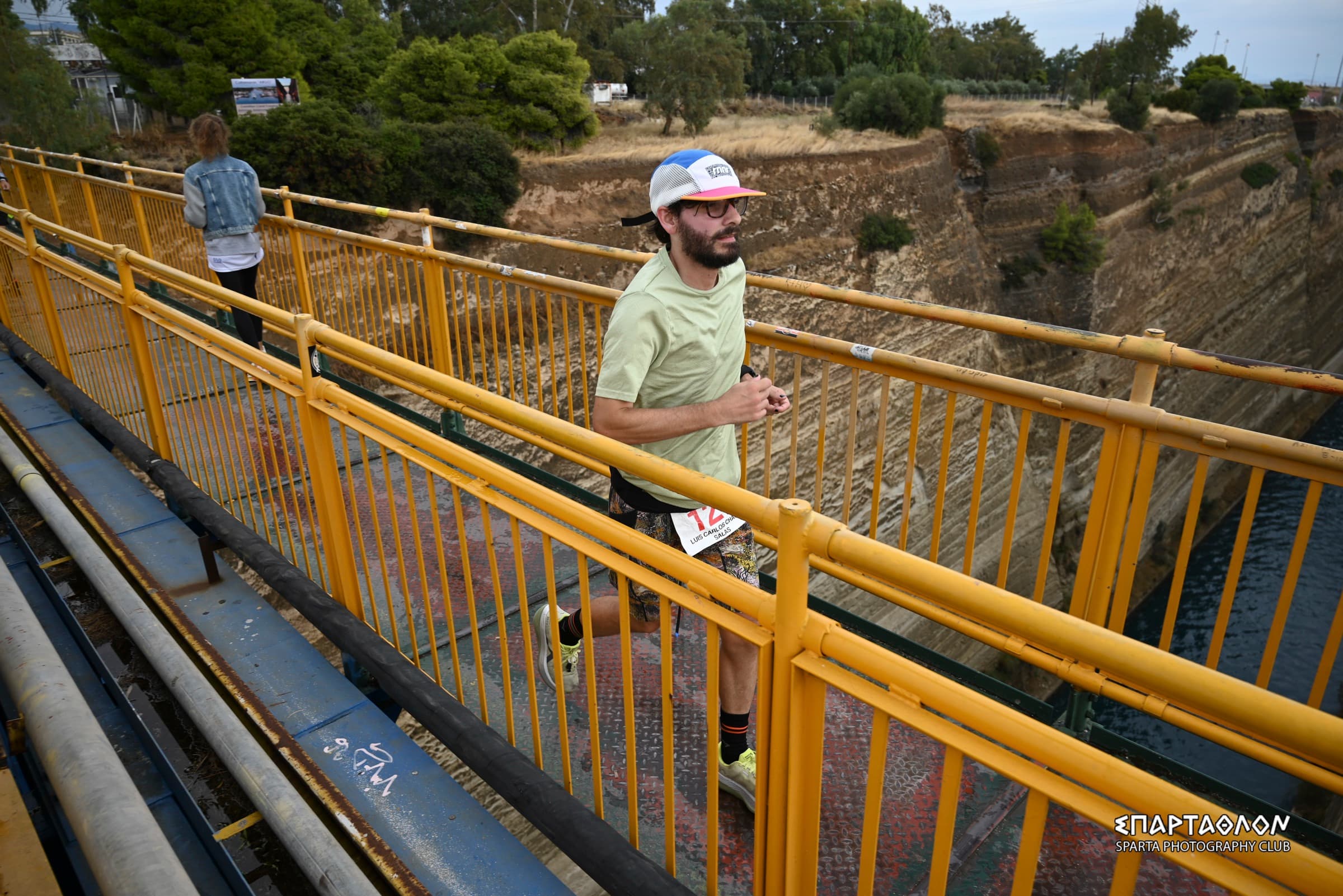El autor cruza corriendo el puente amarillo sobre el canal de Corinto, con el dorsal 127. Abajo se ven las paredes de roca del canal y el agua.