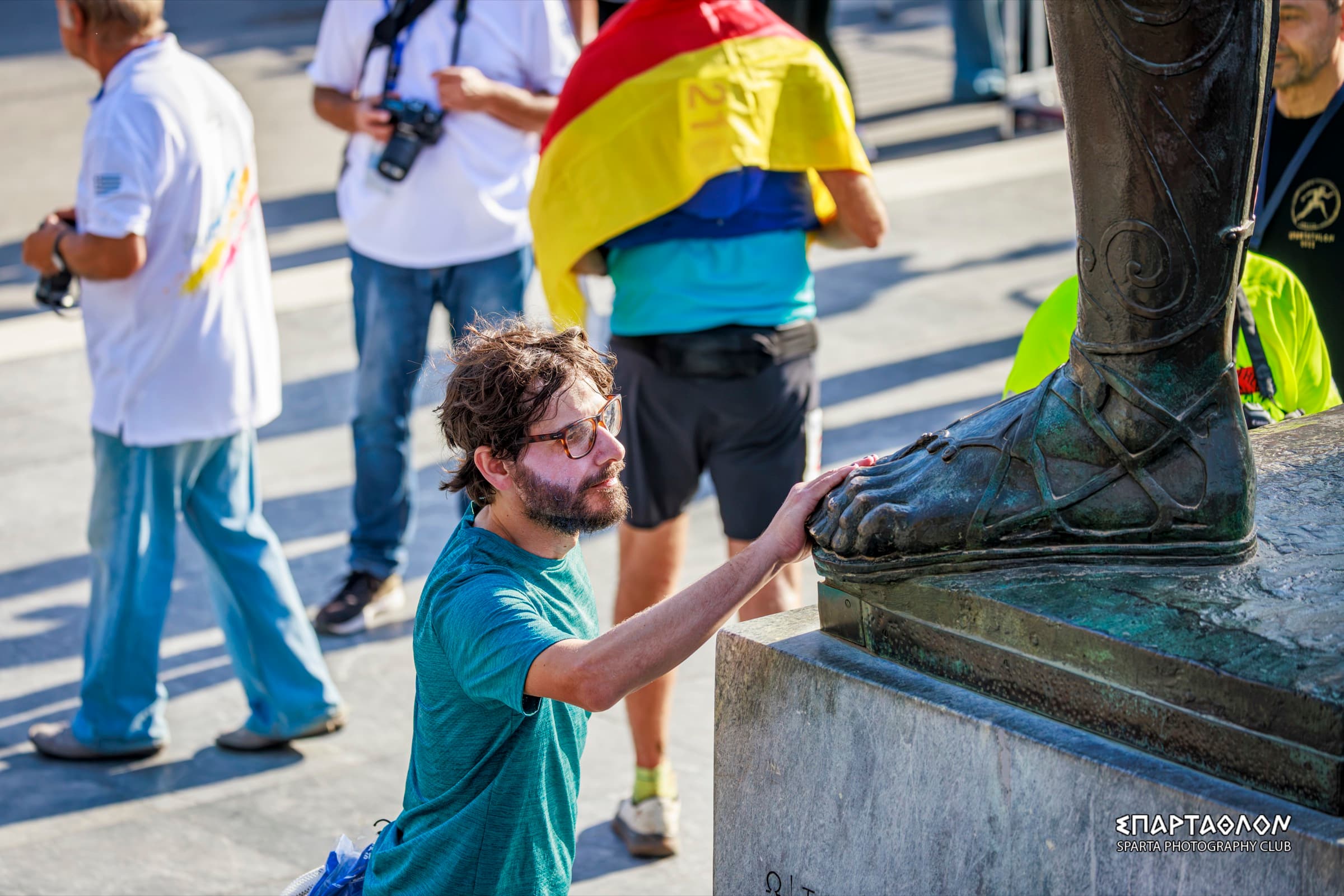 El autor, de rodillas junto a la estatua de Leónidas, pone la mano sobre su pie de bronce. Detrás pasan fotógrafos y un corredor con una bandera.